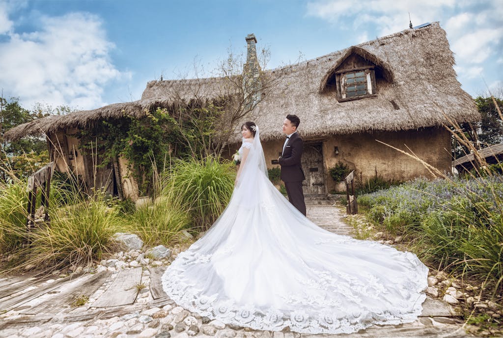 Photo by Lưu Đức Anh Bride and groom pose in elegant attire at a rustic outdoor location with a thatched cottage backdrop.